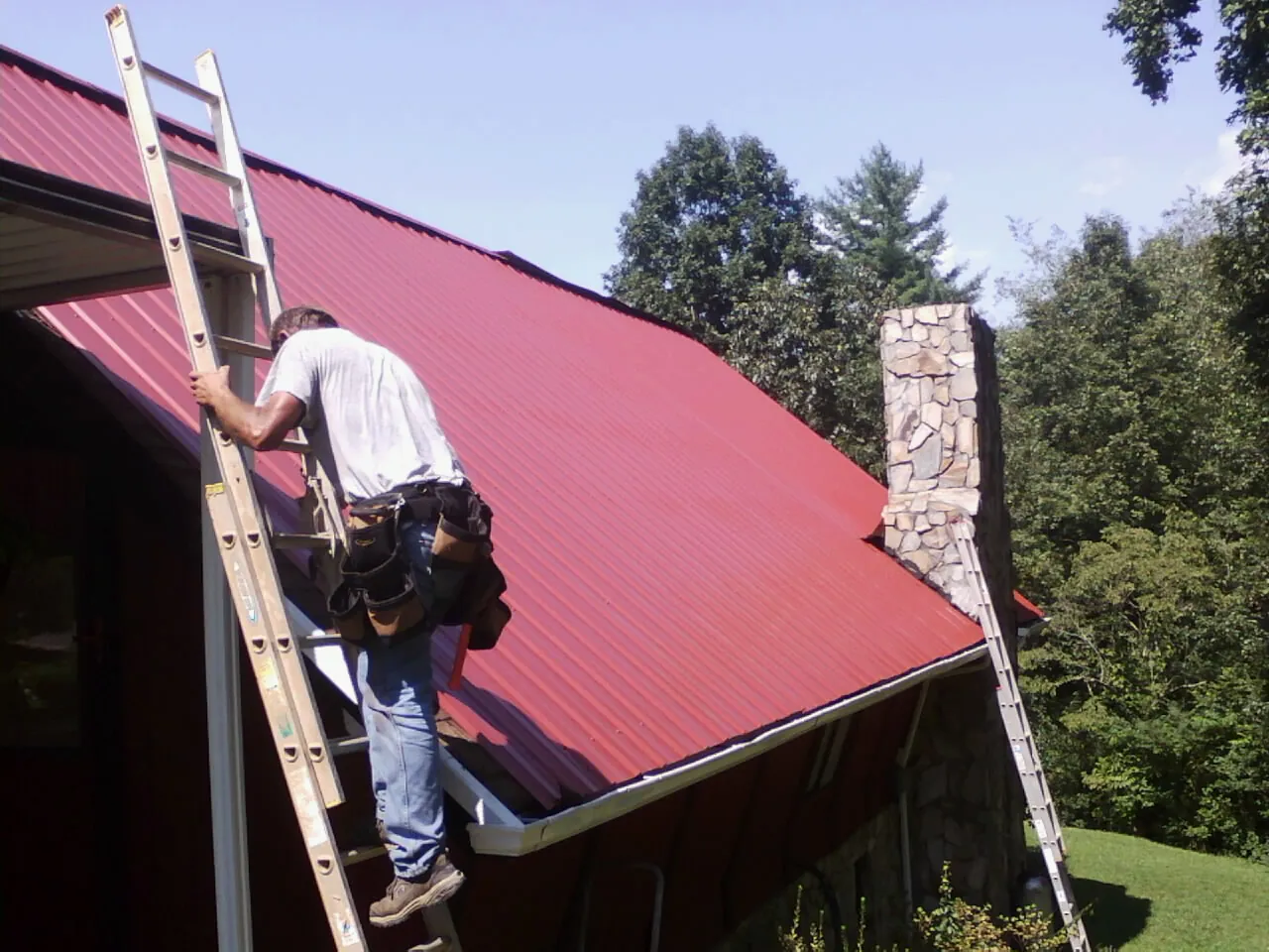 Roofer on a ladder beside a red metal roof and stone chimney — Soffit & Fascia Repair by Roofing Brothers in Lake Butler